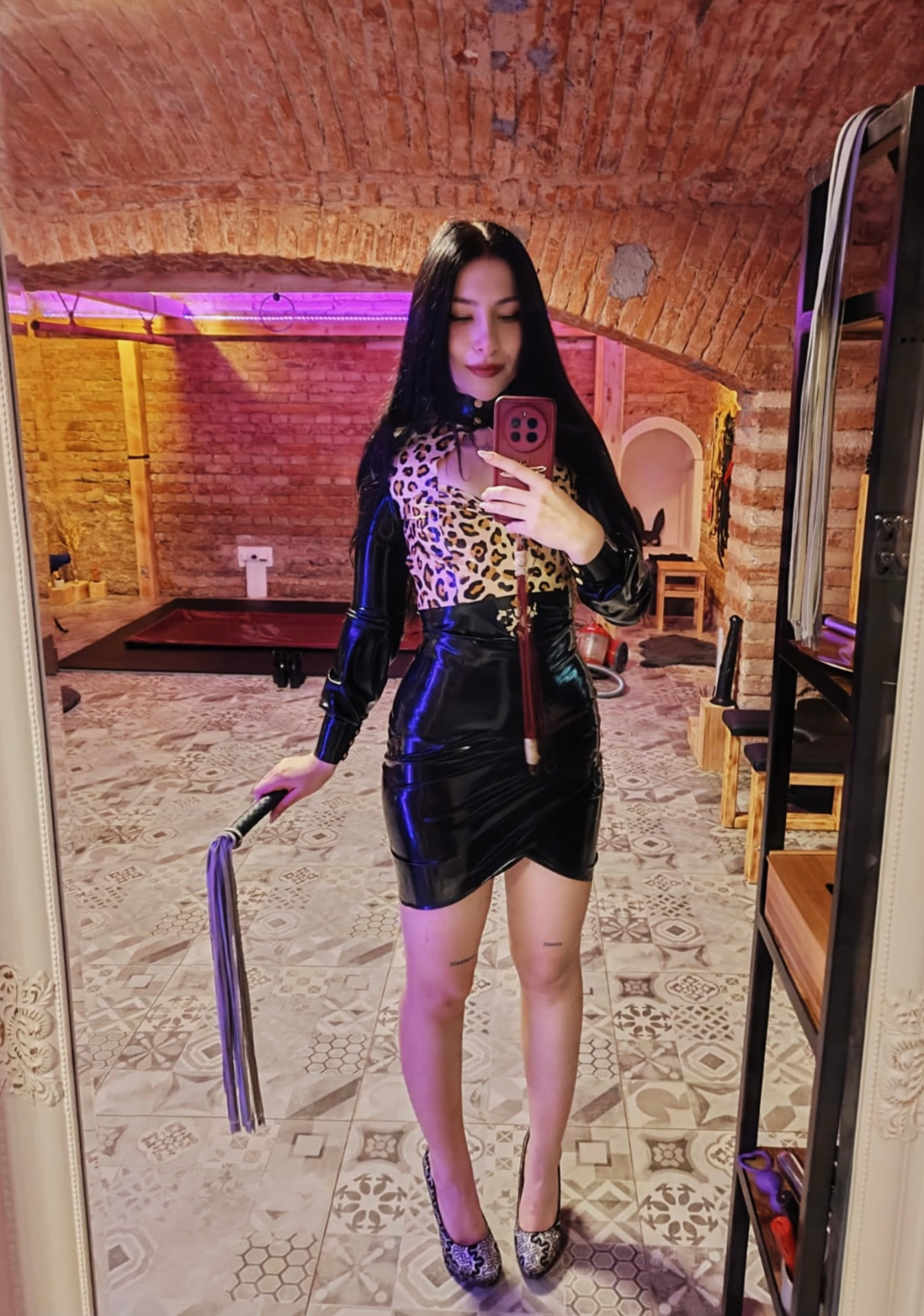 Lady Morgana in black latex holding a flogger in her private studio